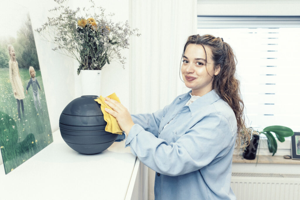 Eine Reinigungskraft in einem blauen Hemd wischt einen runden schwarzen Gegenstand auf einem Regal neben einer Vase und einem Kinderfoto ab.