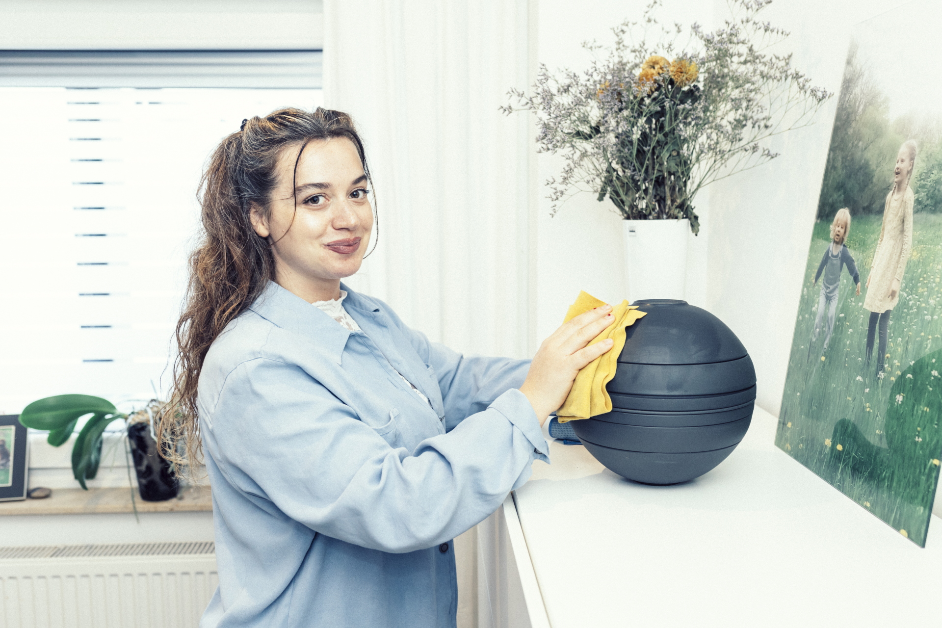 Eine Frau in einem blauen Hemd wischt einen schwarzen Gegenstand auf einer weißen Arbeitsplatte neben einer Vase ab.