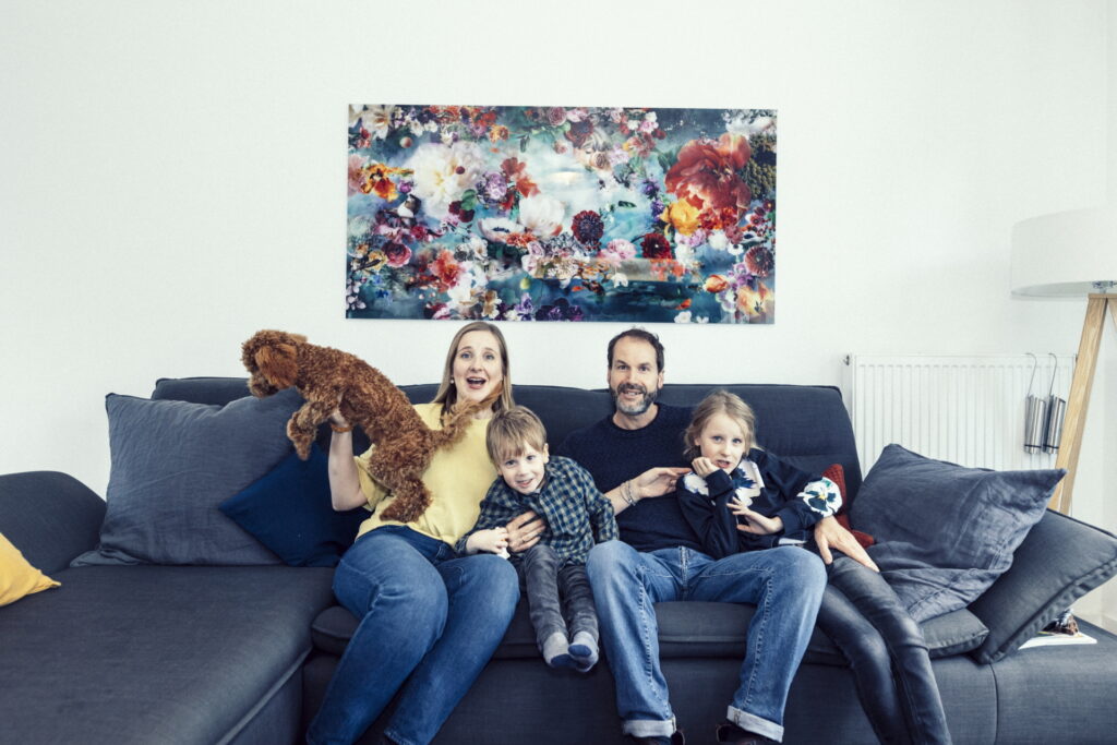 Eine vierköpfige Familie mit Hund sitzt auf einem grauen Sofa und verbringt Zeit im Wohnzimmer, das mit bunten Wandbildern geschmückt ist.
