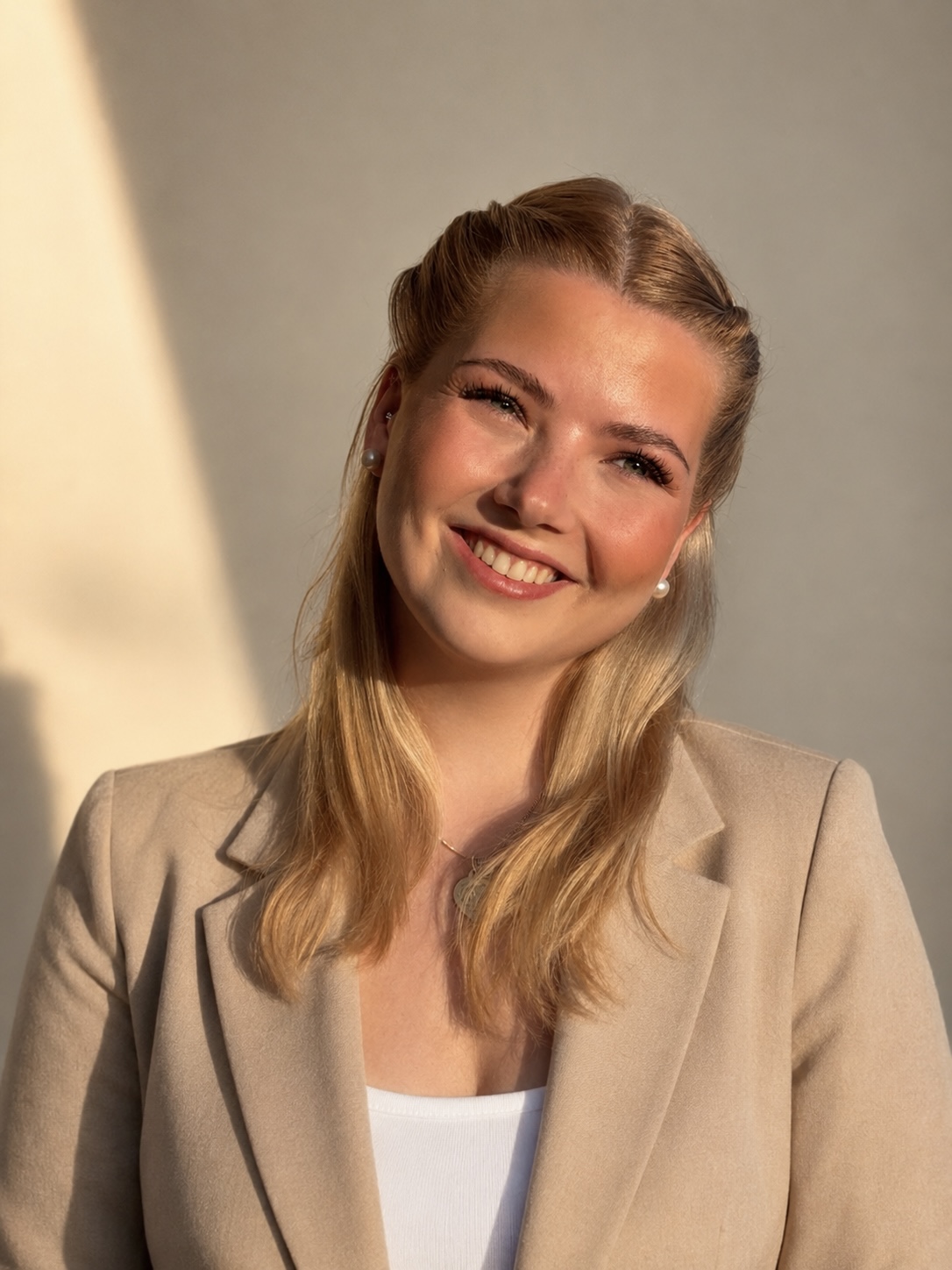 Eine junge Frau mit blonden Haaren in einem beigen Blazer lächelt vor einem schlichten Hintergrund im Sonnenlicht in die Kamera.