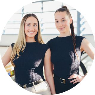 Zwei Frauen stehen drinnen nebeneinander, tragen ärmellose schwarze Oberteile und lächeln in die Kamera.