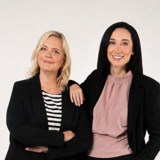 Zwei Frauen in Businesskleidung stehen nebeneinander vor einem schlichten Hintergrund, eine blond, eine dunkelhaarig.